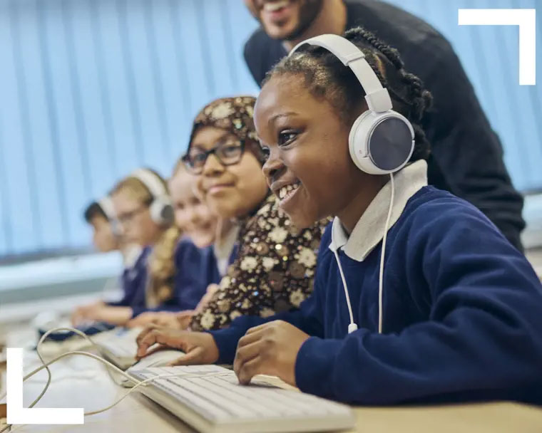 Teacher assisting smiling key stage 1 pupil who wears headphones as they work on a desktop computer